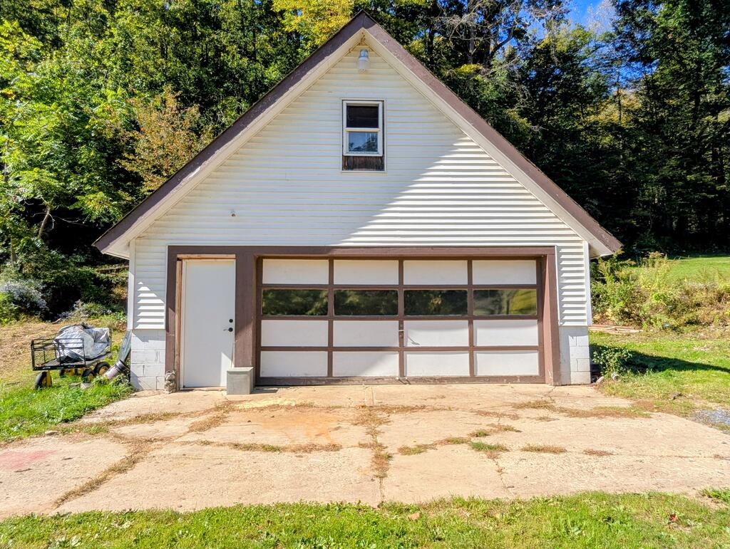 Detached white garage with a steep gable roof and wide paneled door, surrounded by trees.