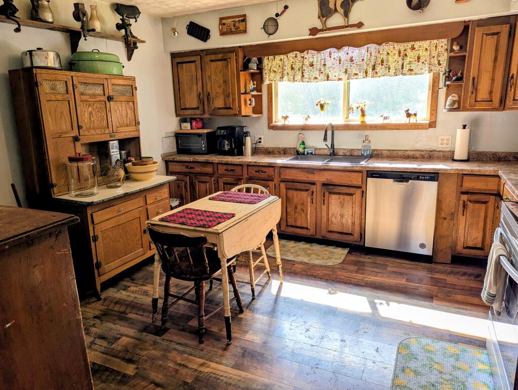 Rustic kitchen with wood cabinets, a farmhouse-style table, and a double sink beneath a bright window.