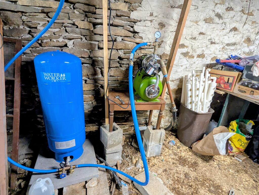 Basement water system with a blue pressure tank, green pump, and exposed piping against stone walls.