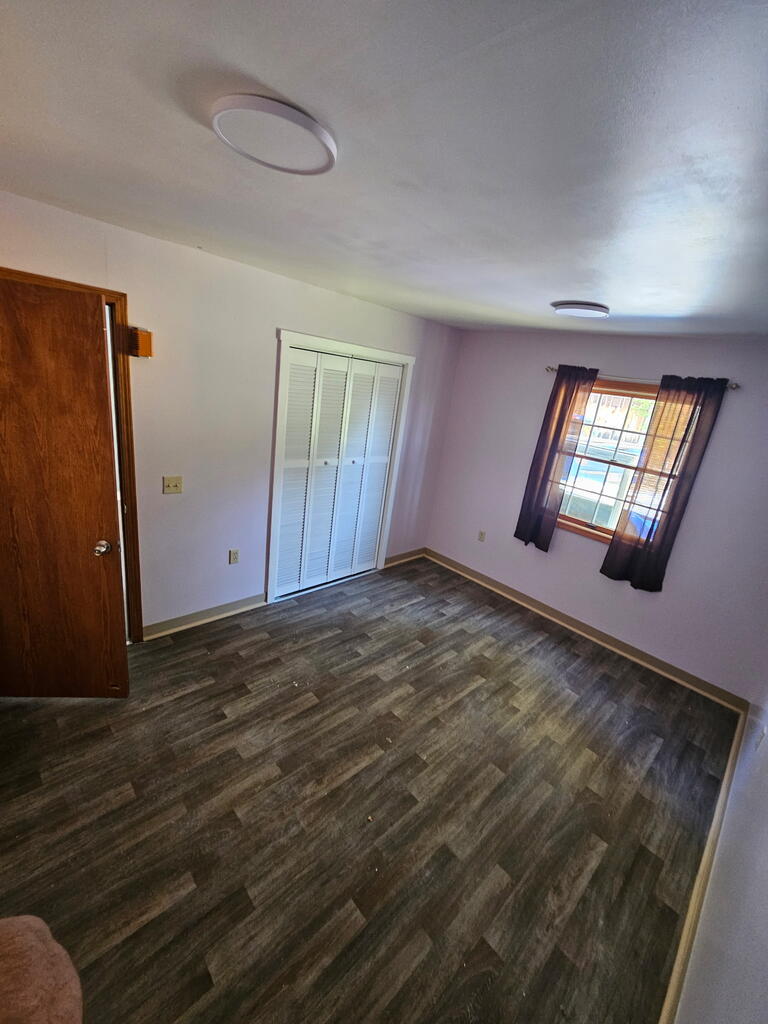 Empty bedroom with a louvered closet, dark plank flooring, and a curtained window.