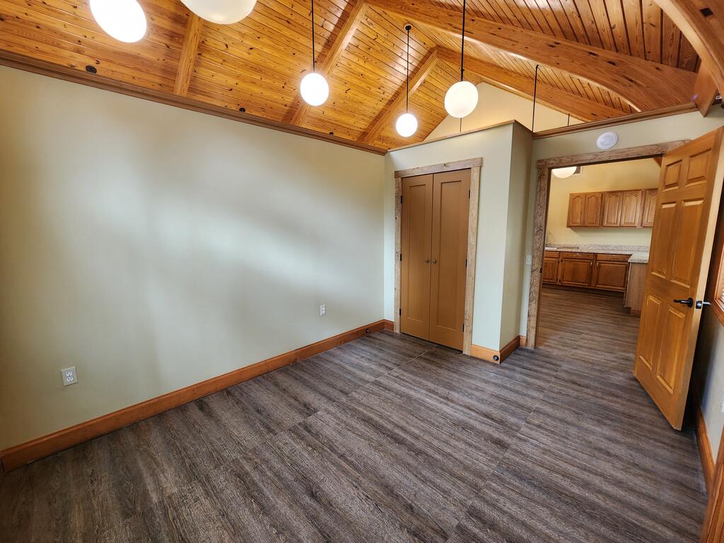 Empty room with a vaulted wood ceiling, gray flooring, and an open doorway to the kitchen.