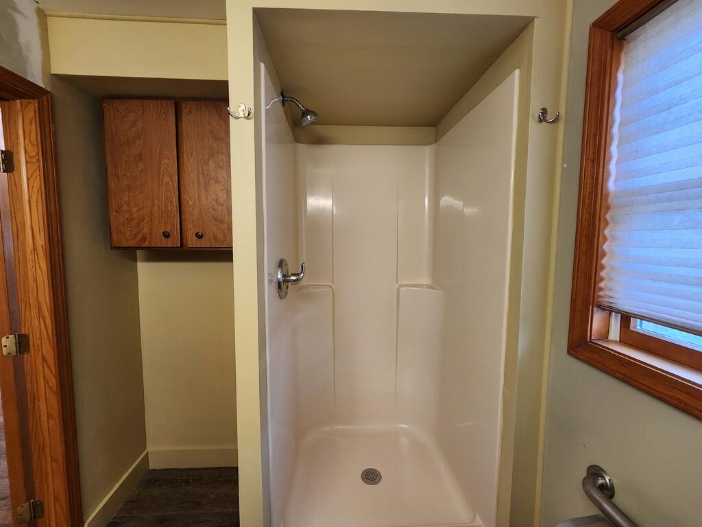 Bathroom shower stall with a built-in seat, wooden cabinets, and a window with a blind.