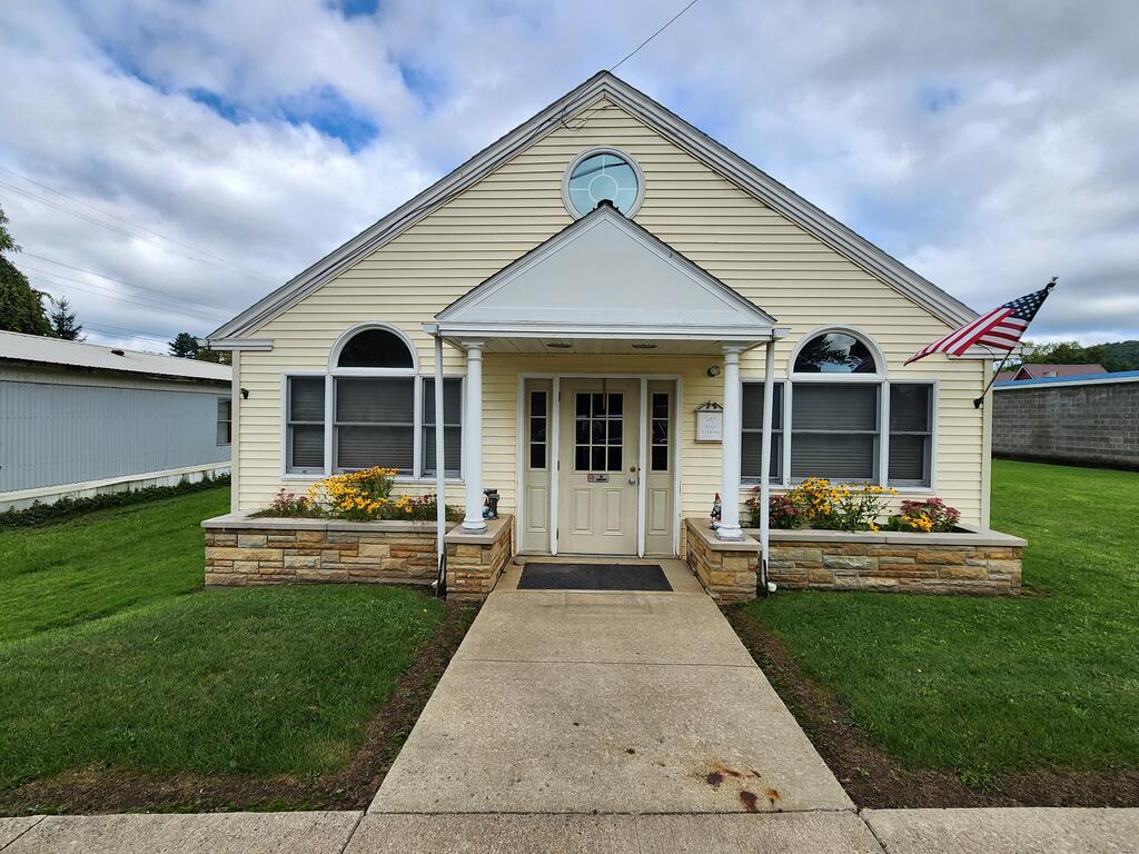 Cream gabled building with a columned entrance, flower beds, and an American flag.
