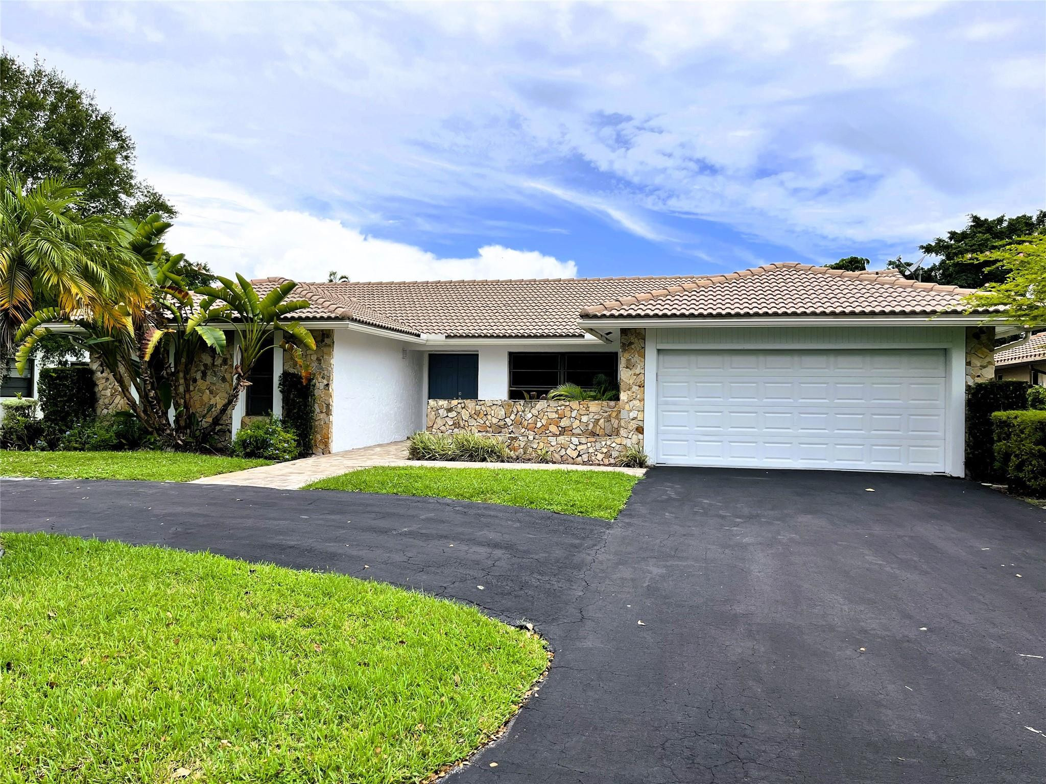 Single-story home with stone accents, a white garage door, tropical landscaping, and an asphalt driveway.