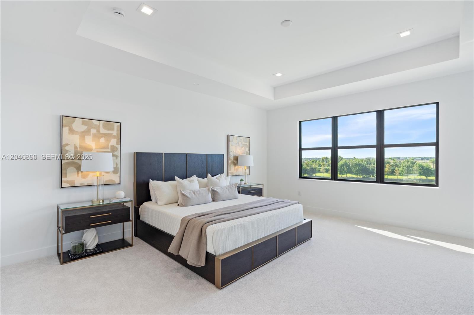 Staged bedroom with a dark bed, white bedding, and large windows overlooking green trees.