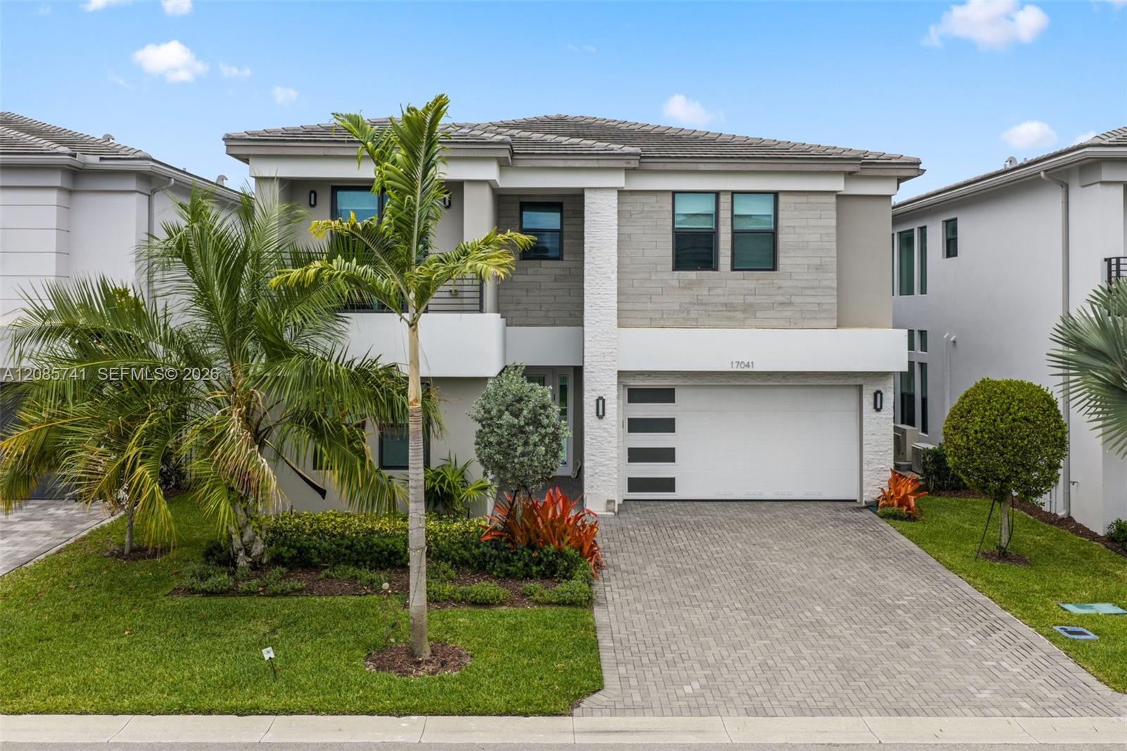 Modern two-story home with a gray stone facade, white garage, paved driveway, and tropical palms.