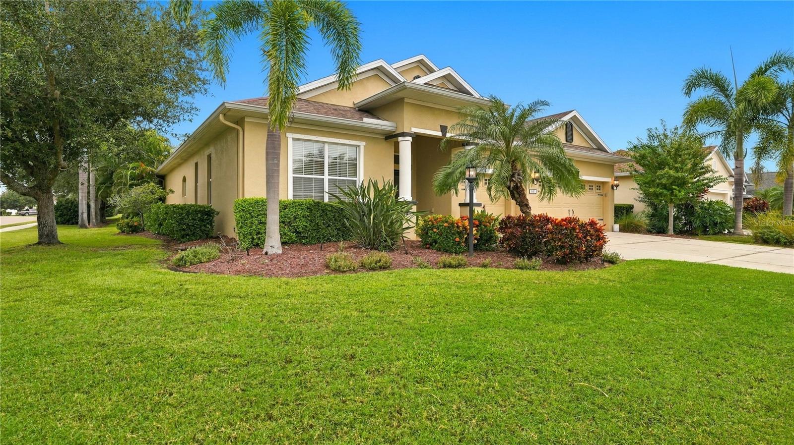 Tan stucco home with an attached garage, palm trees, colorful landscaping, and a broad green lawn.
