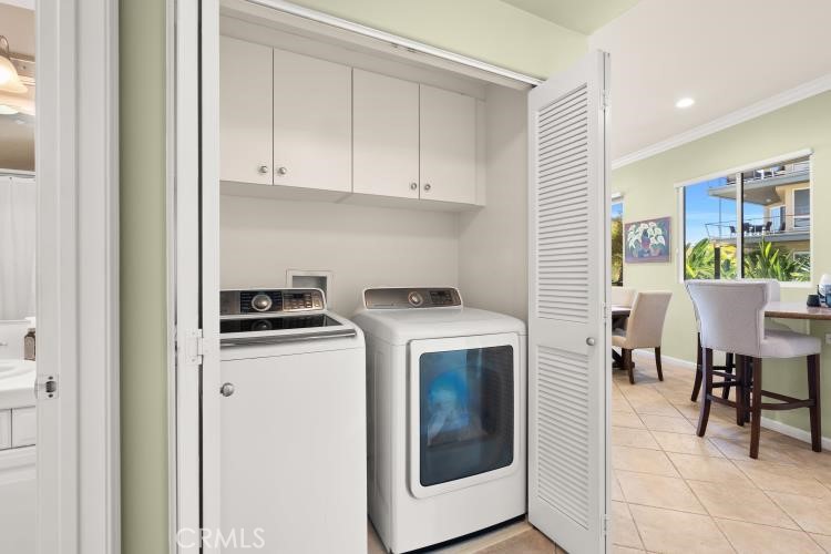 Laundry closet with washer and dryer beneath overhead cabinets, beside a bright dining area.