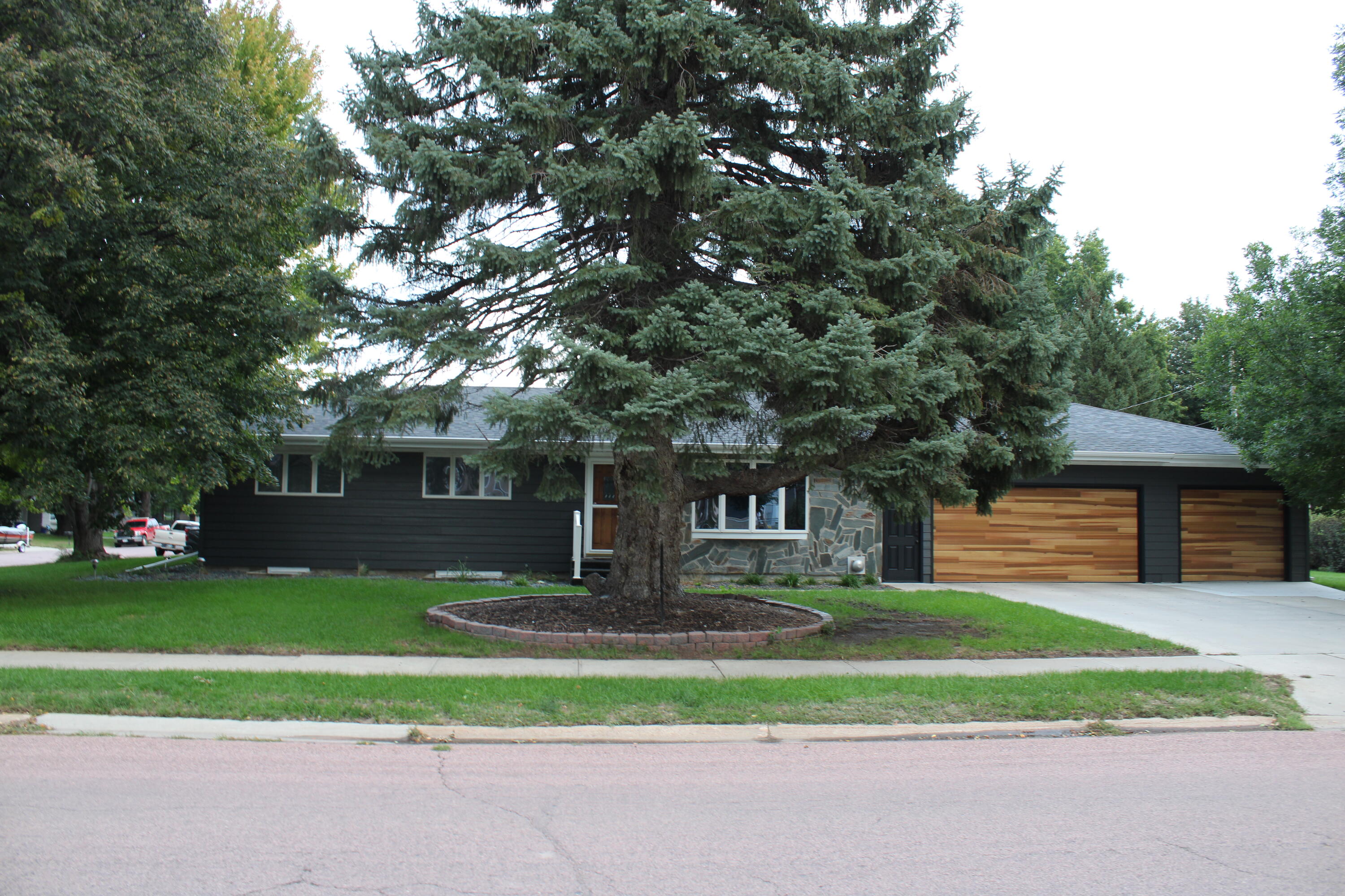 Single-story dark-sided house with stone facade and wood-paneled garage doors, partly obscured by a large spruce.