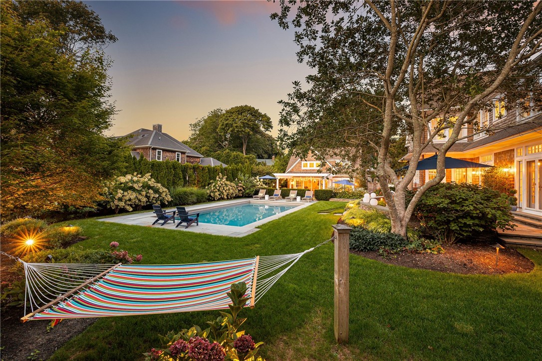 Backyard pool and striped hammock beside a warmly lit house at dusk.