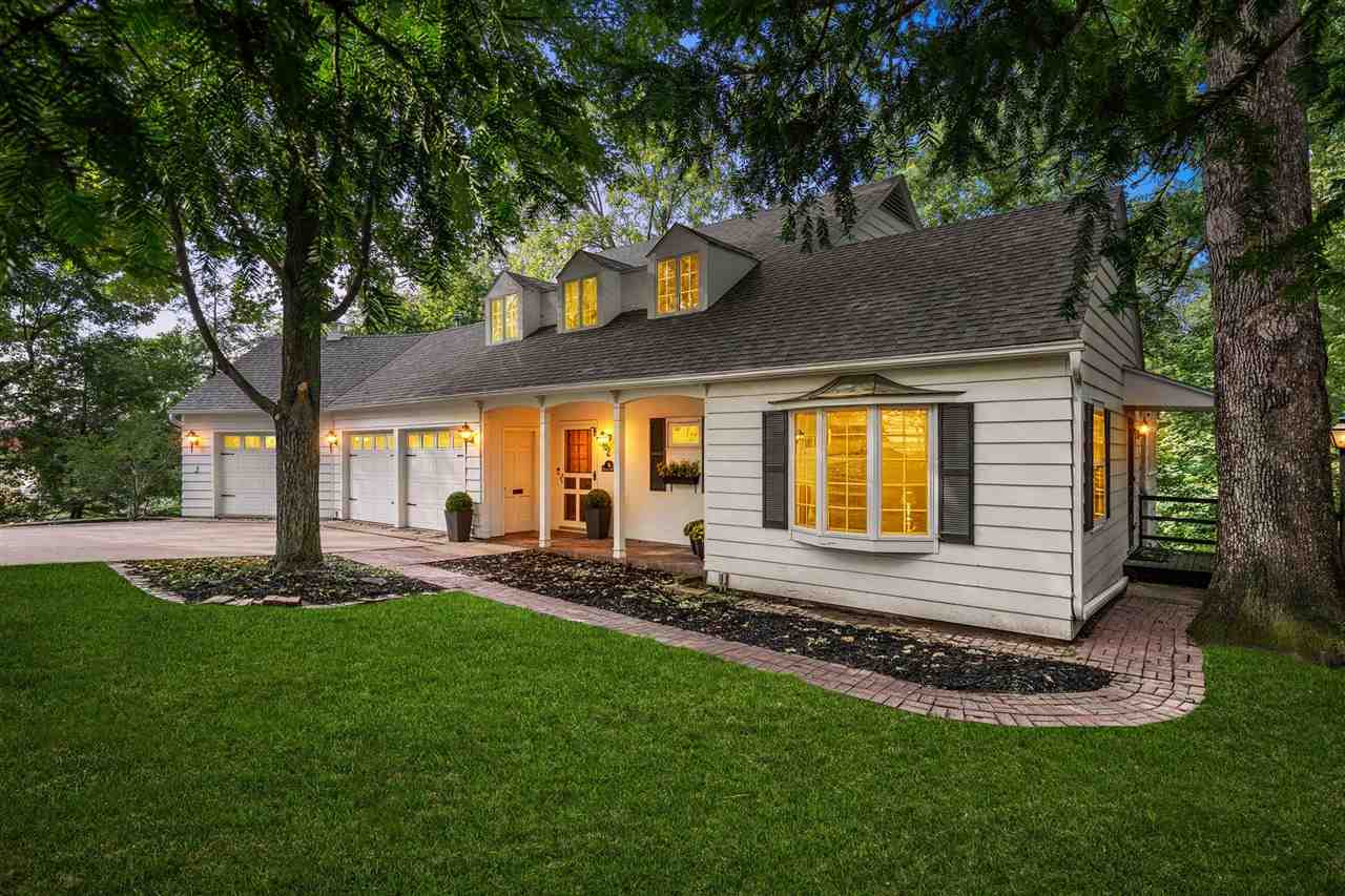 White-sided house with lit dormers and three-car garage, framed by mature trees and a curved brick walkway.
