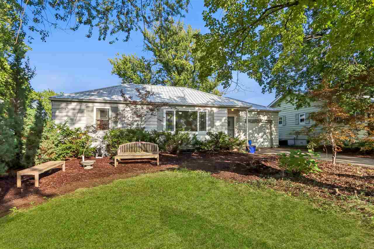 Single-story gray home with a metal roof, framed by mature trees and a landscaped yard with benches.