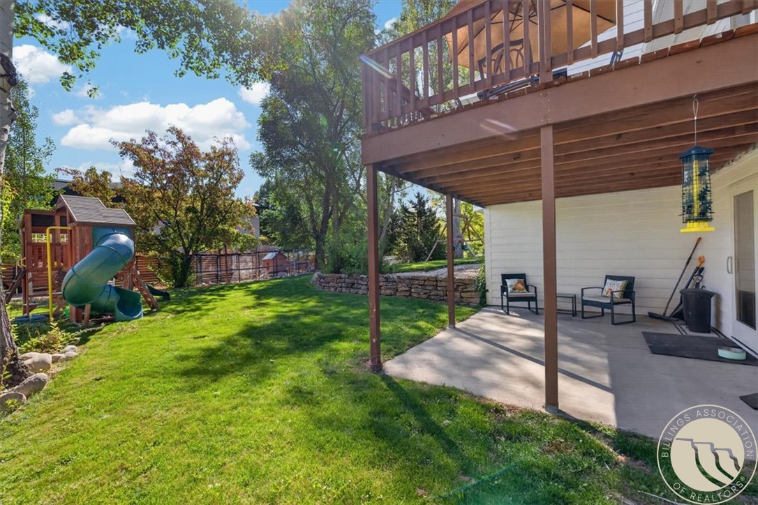 Backyard patio beneath a raised wooden deck, overlooking a lawn and children's playset.