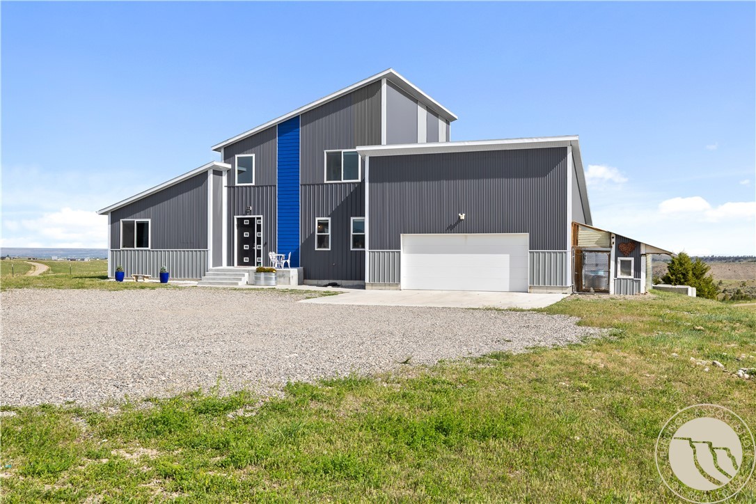 Modern gray metal-sided home with a blue accent panel and attached garage beside a broad gravel driveway.