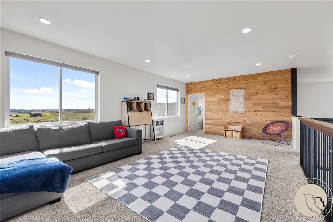 Upstairs living area with a gray sectional, checkerboard rug, wood-plank accent wall, and windows overlooking open land.