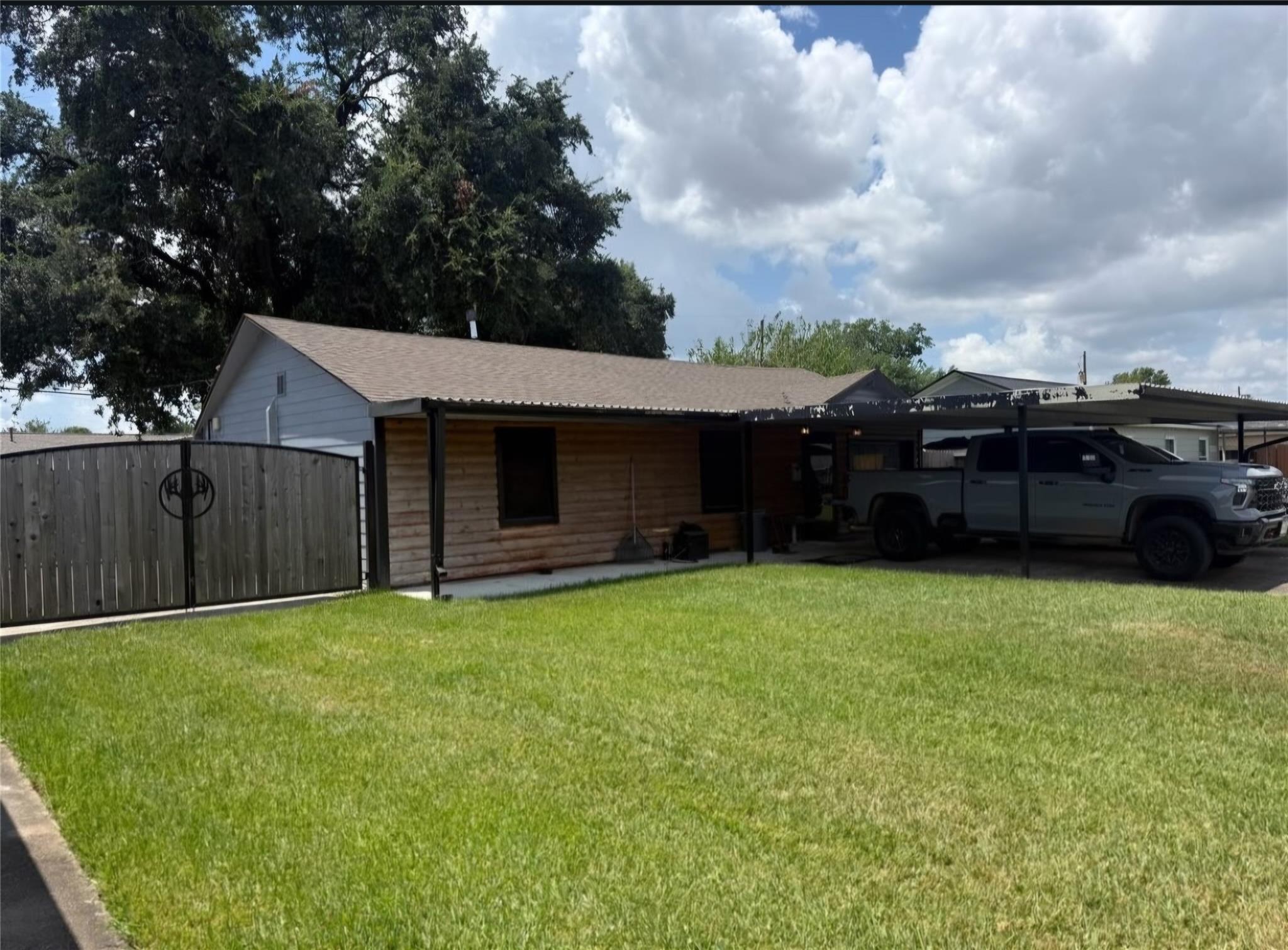 Single-story wood-sided home with a covered carport, pickup truck, fenced yard, and broad green lawn.