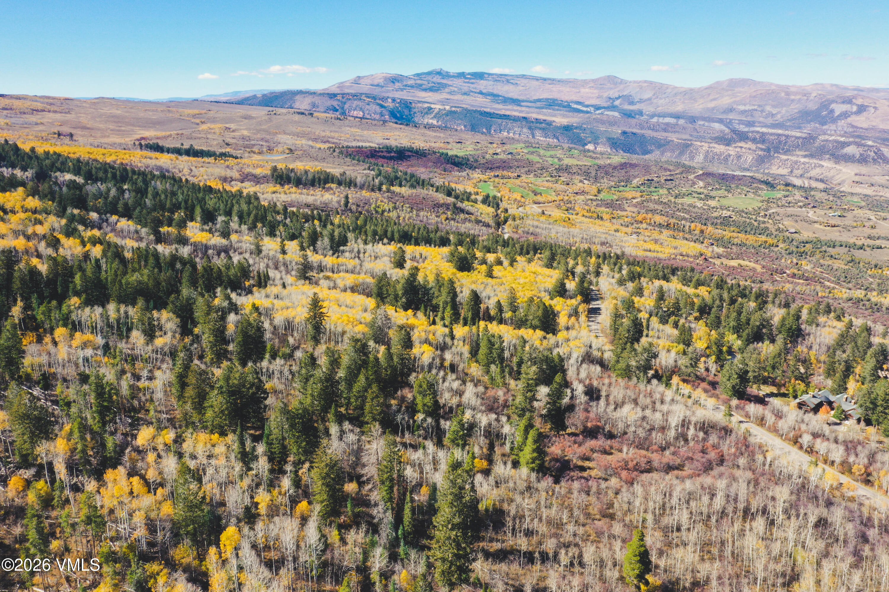 Aerial view of autumn-colored forest and mountain ranges, with an access road winding past scattered homes.