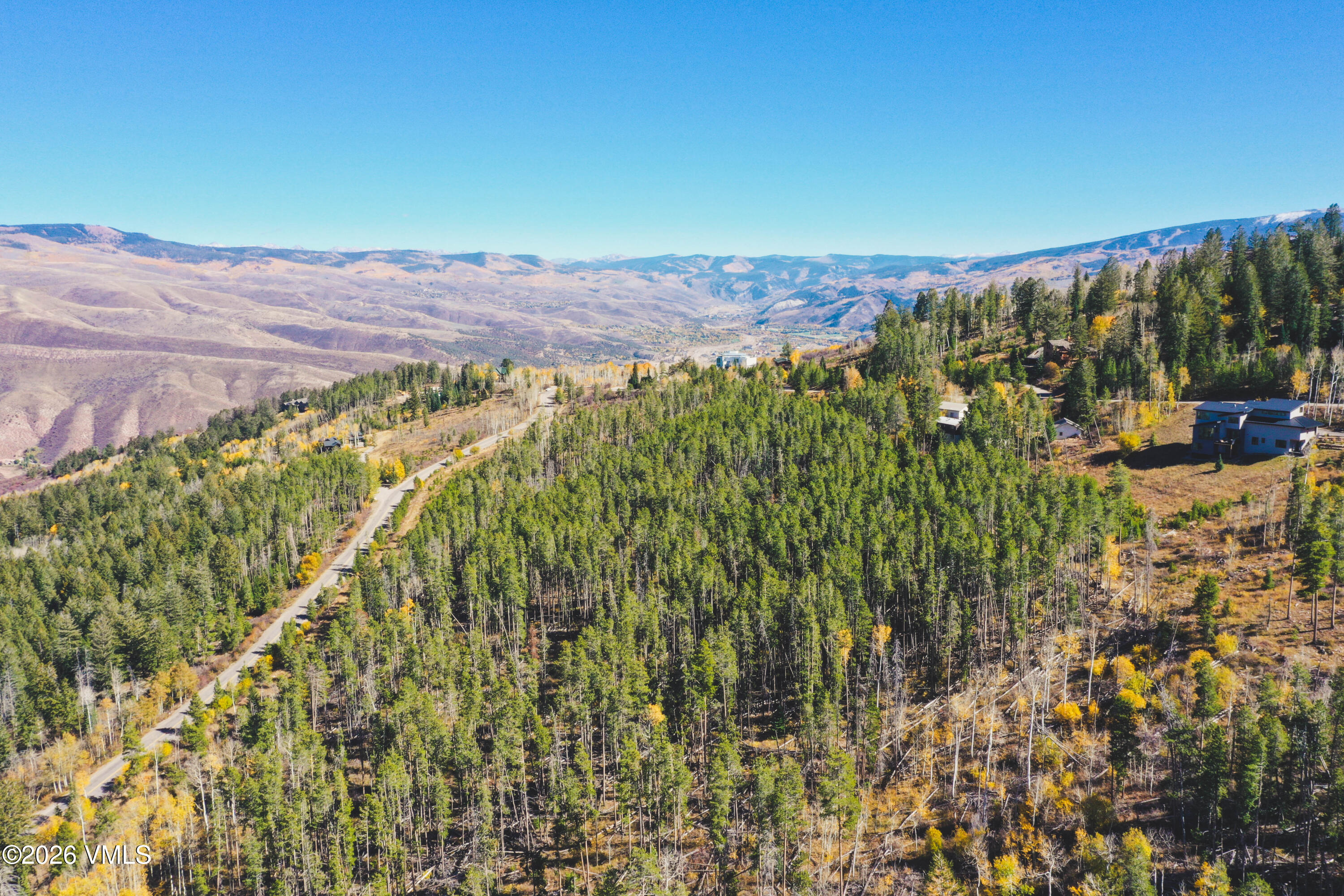 Aerial view of a wooded hillside lot, winding road, nearby homes, and distant mountain ranges.