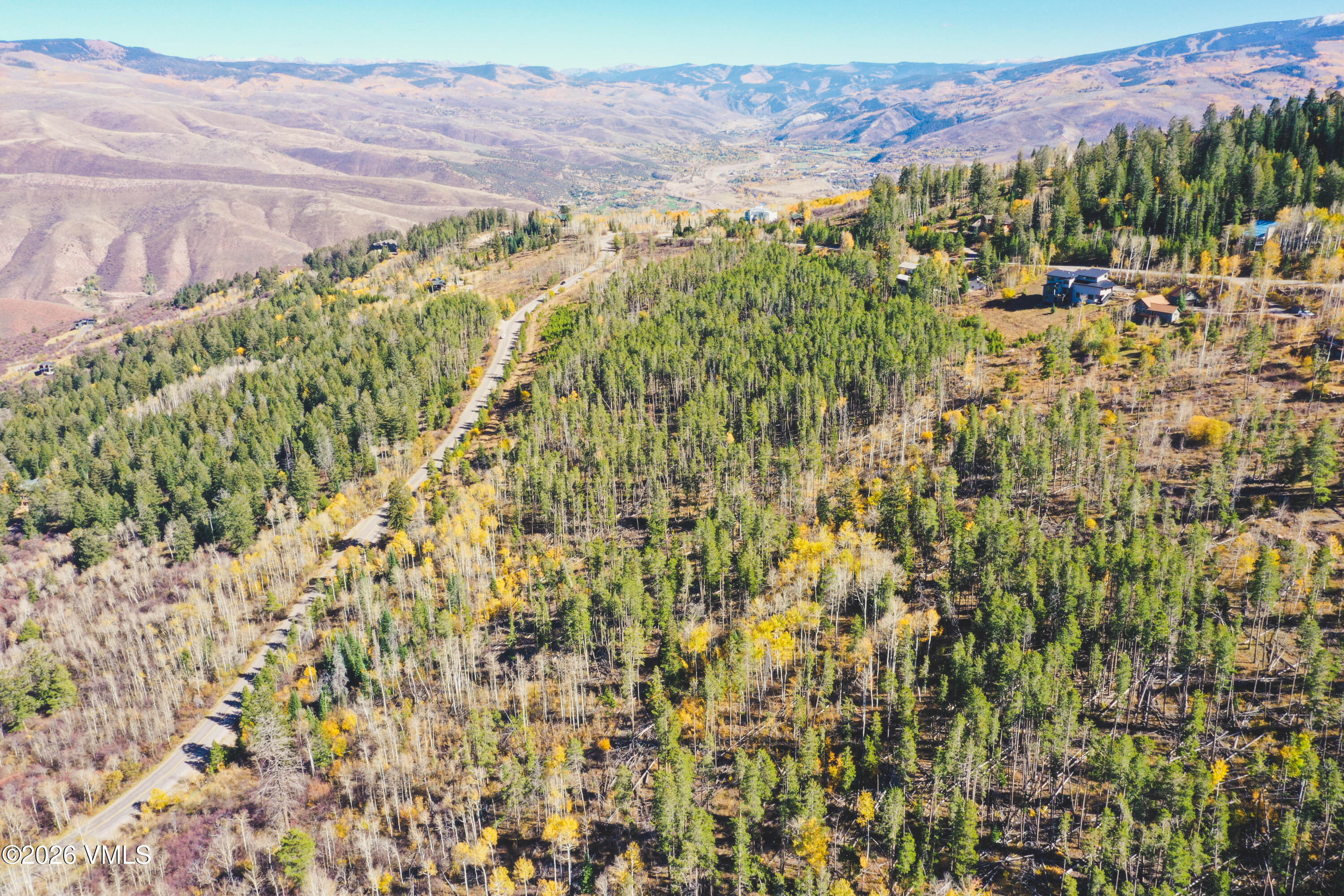 Aerial view of wooded hillside land, a winding road, nearby homes, and distant mountains.