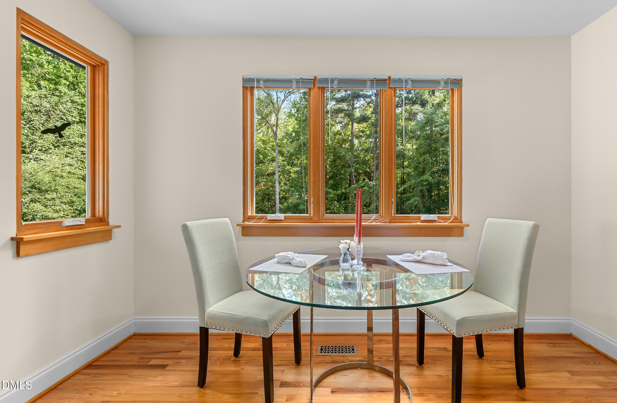 Dining area with a glass-top table and two chairs beside large windows overlooking trees.