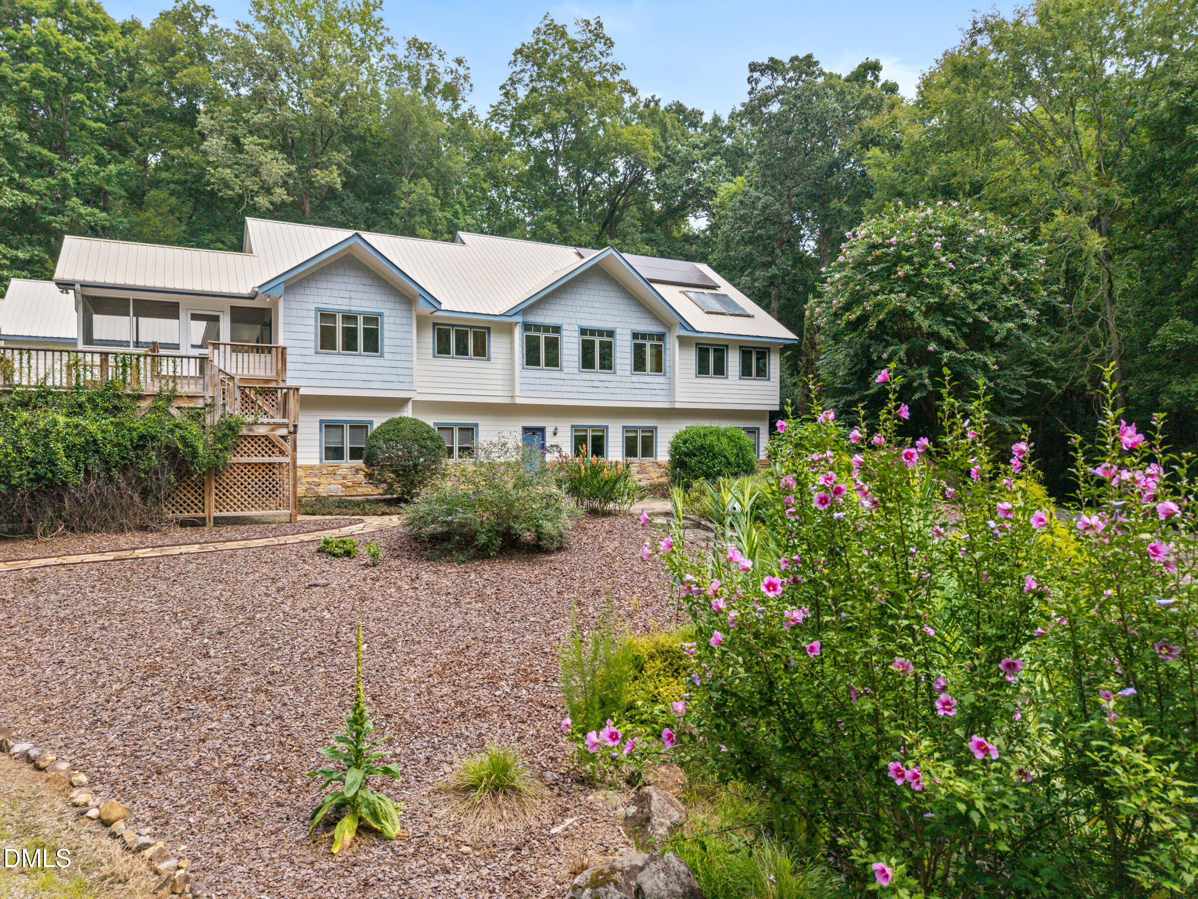 House with a metal roof and raised deck, set behind a garden of pink flowers and mature trees.