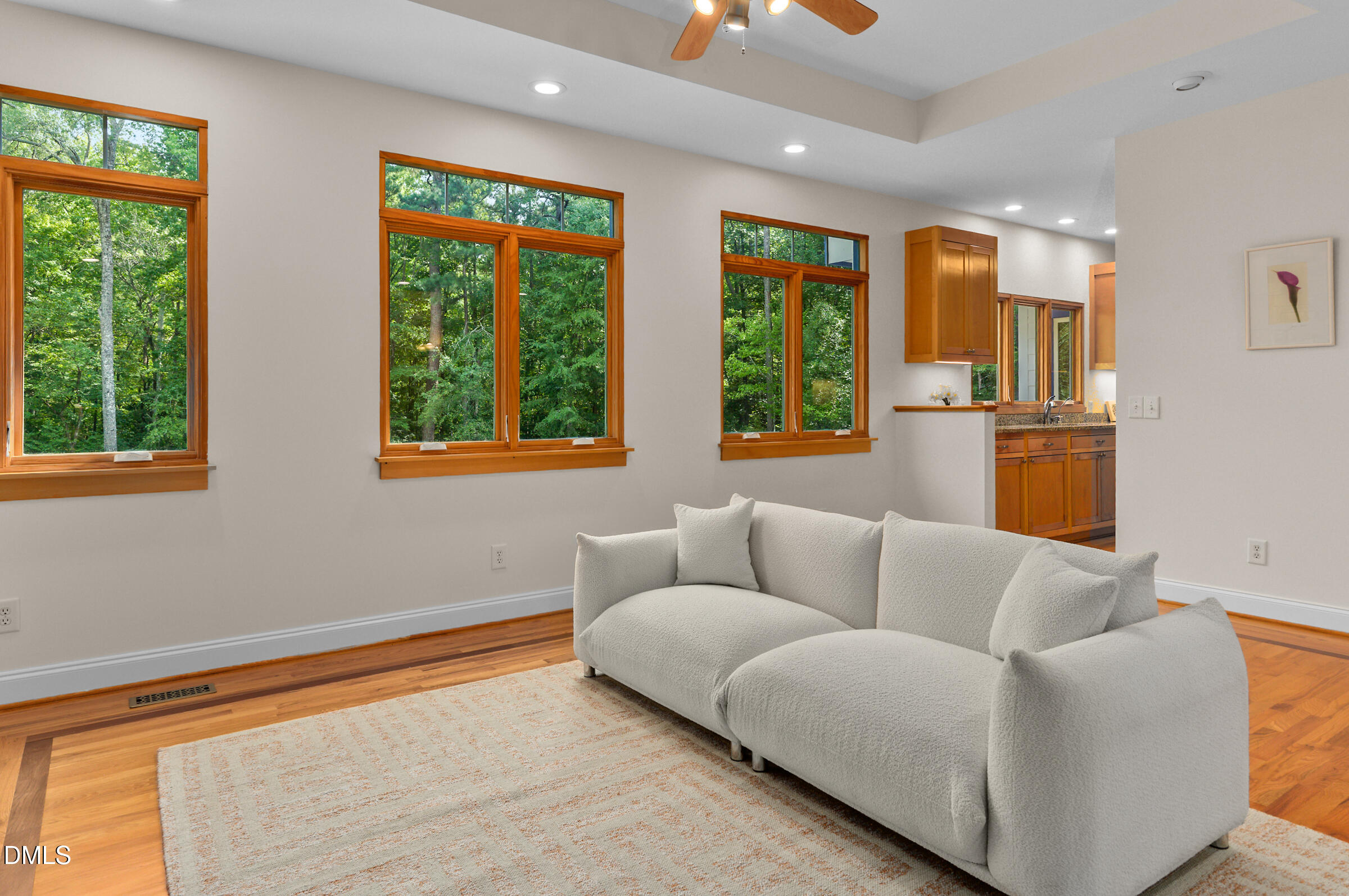Living room with a white sectional, hardwood floors, and wood-trimmed windows overlooking trees.