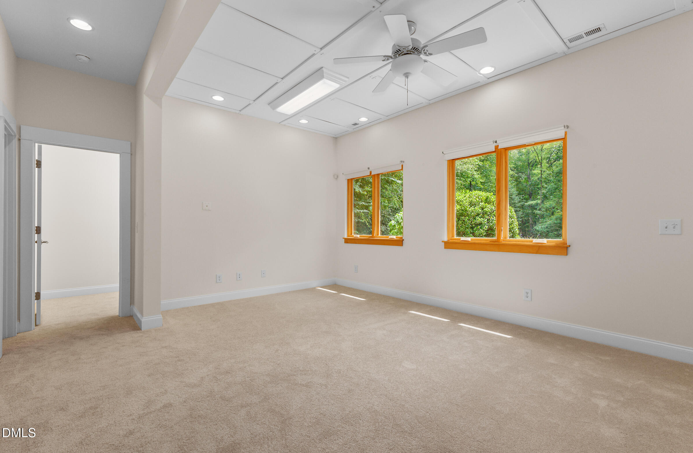 Empty carpeted room with a ceiling fan and wood-trimmed windows overlooking trees.