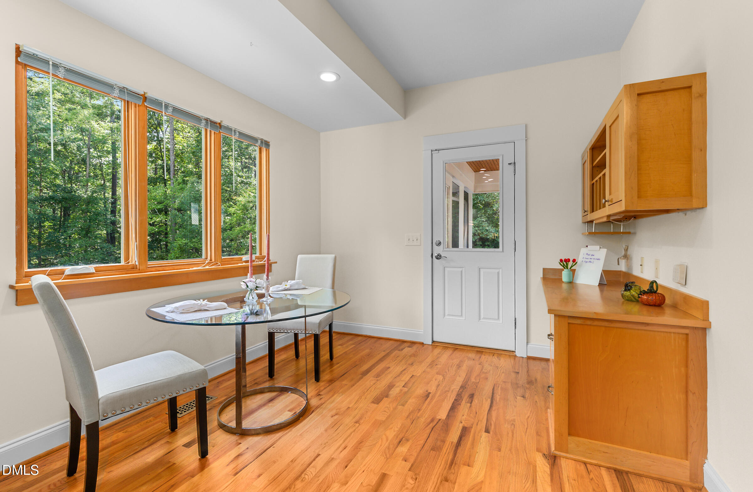 Dining area with an oval glass table, hardwood floors, built-in cabinetry, and large windows overlooking trees.