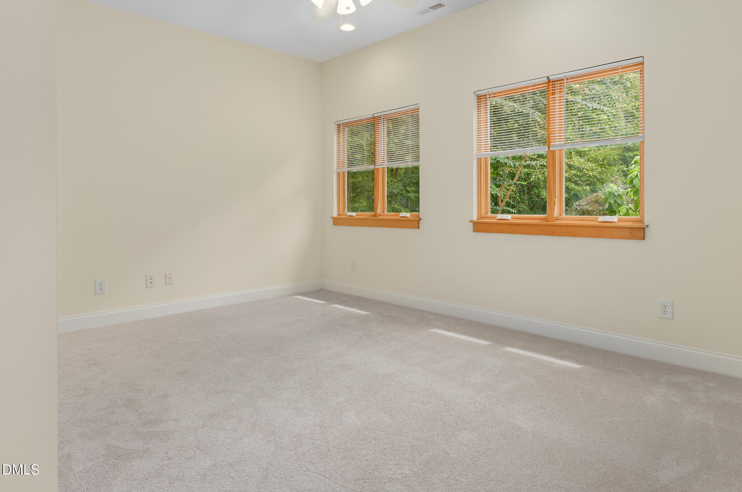 Empty carpeted room with two sets of wood-trimmed windows overlooking leafy trees.