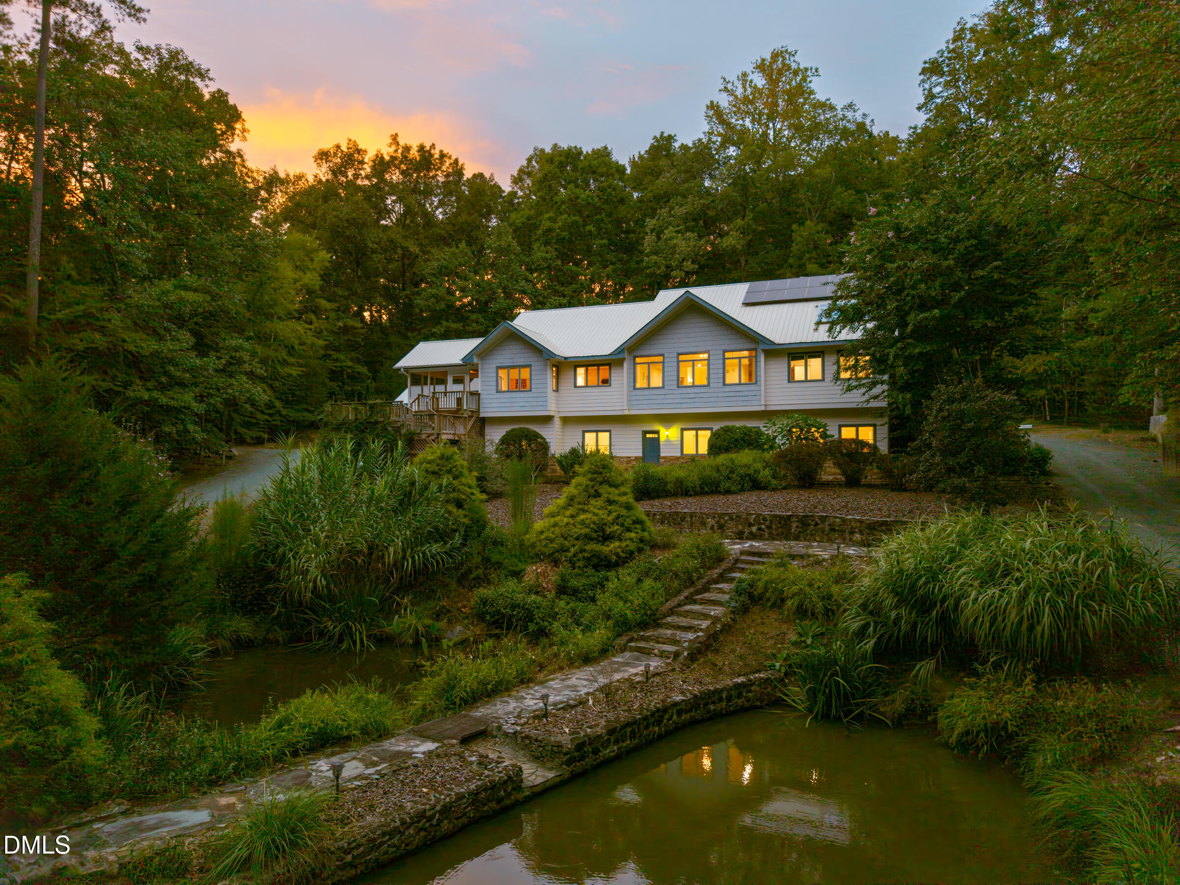 Two-story gray home with solar panels, glowing windows, and a stone path beside a pond at dusk.