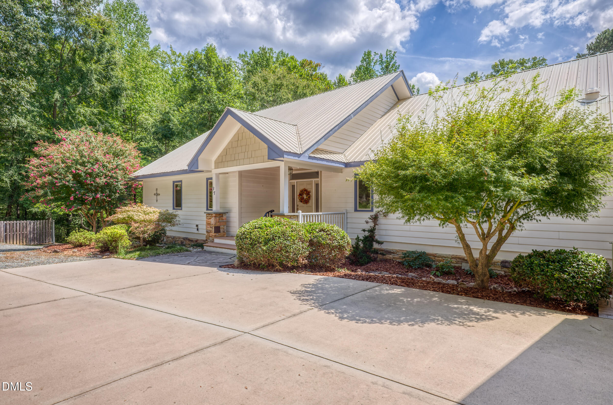 Light-colored home with a metal roof, covered entry porch, flowering shrubs, and a wide concrete driveway.