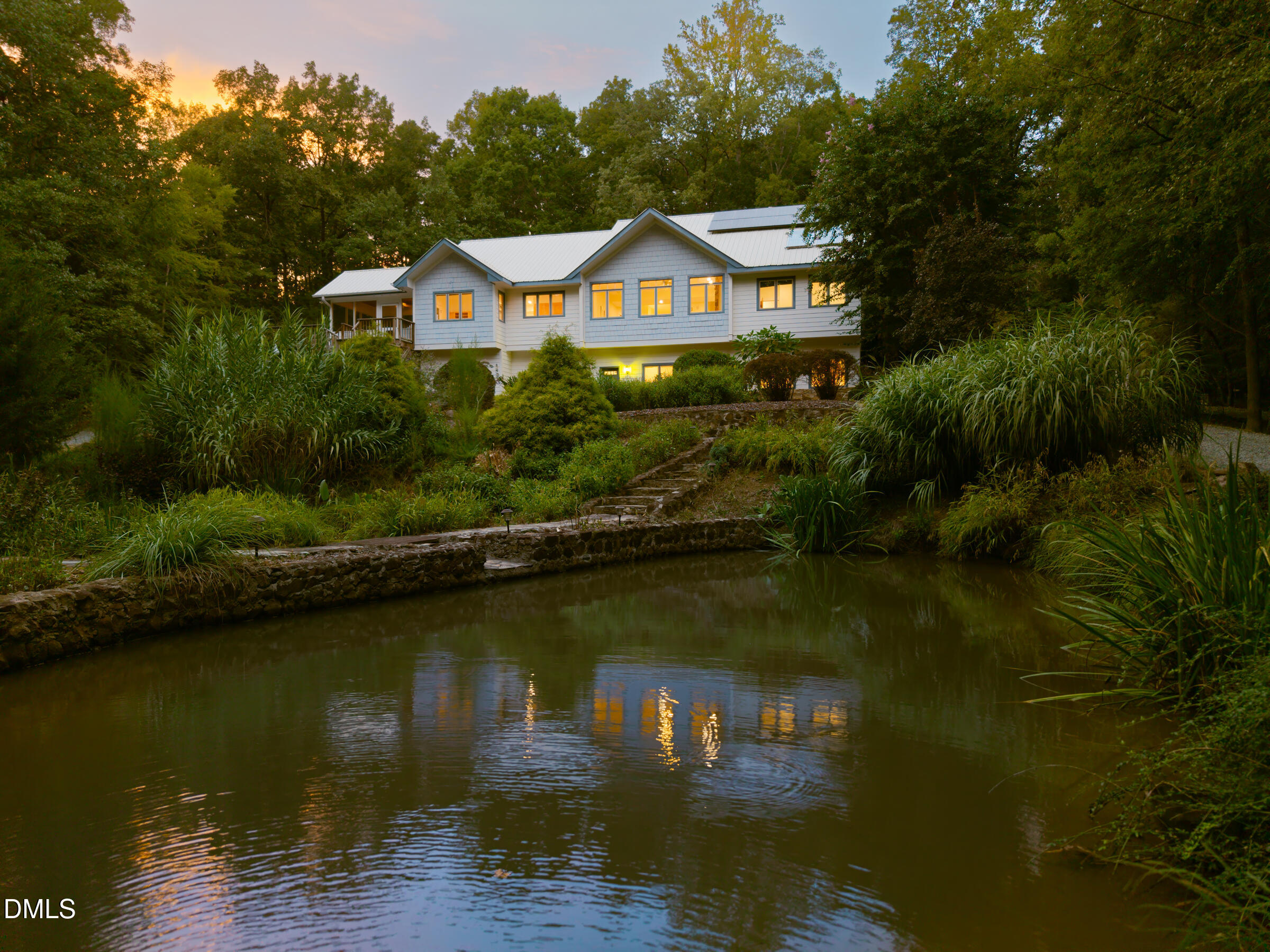 Modern home among trees overlooking a landscaped pond, with warm window lights reflected in the water.