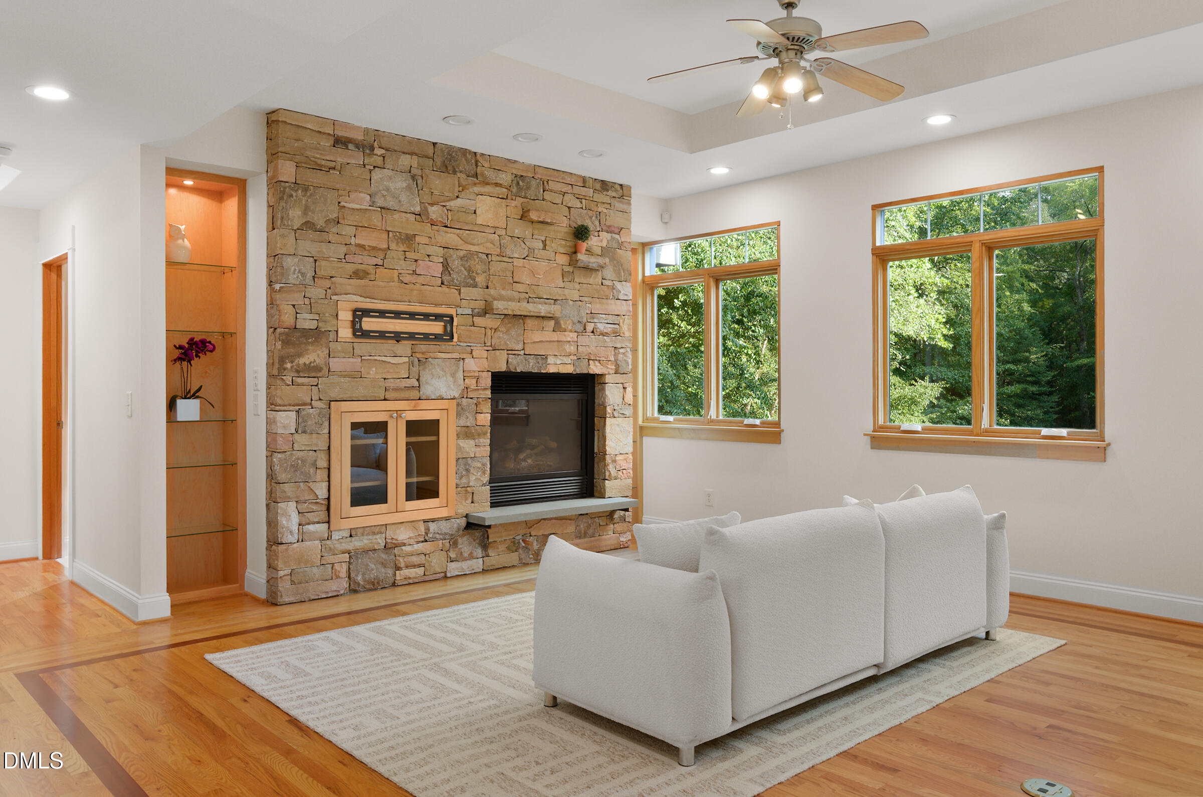 Living room with a floor-to-ceiling stone fireplace, white sofa, hardwood floors, and windows overlooking trees.