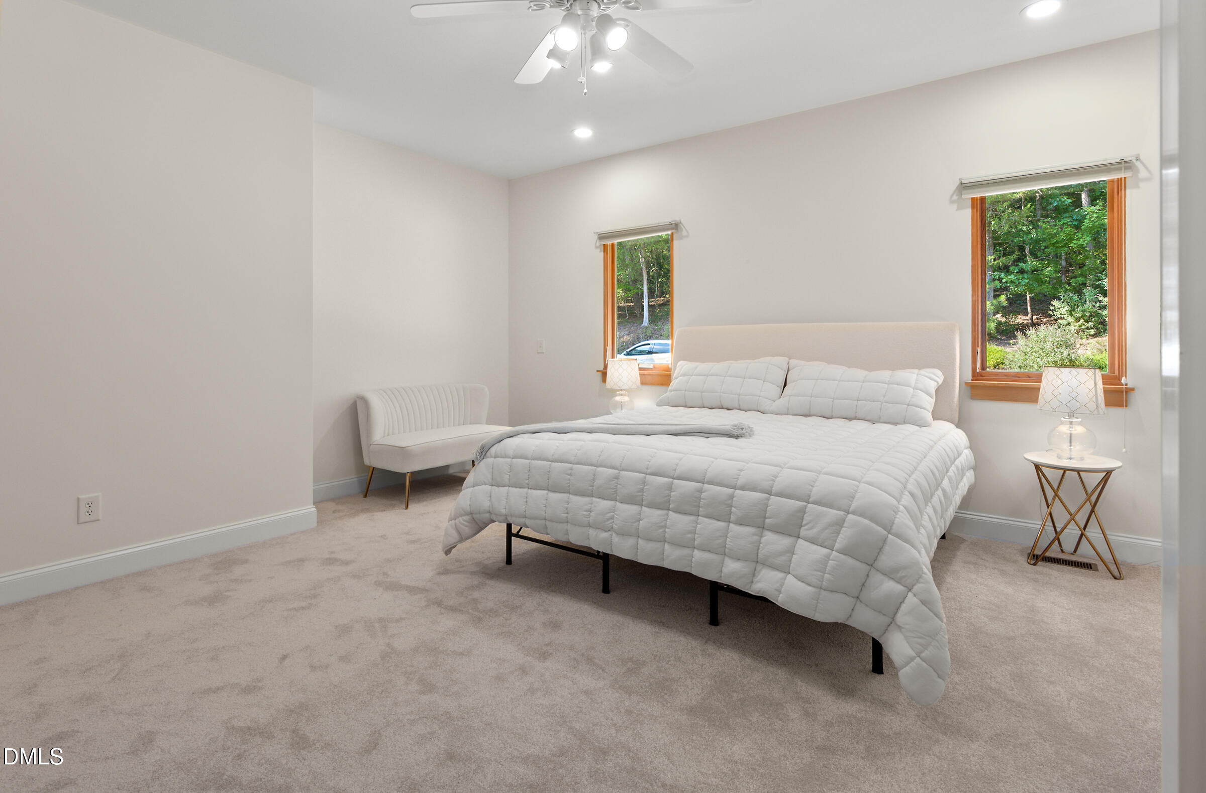 Furnished bedroom with a white bed, cream bench, and two windows overlooking trees.