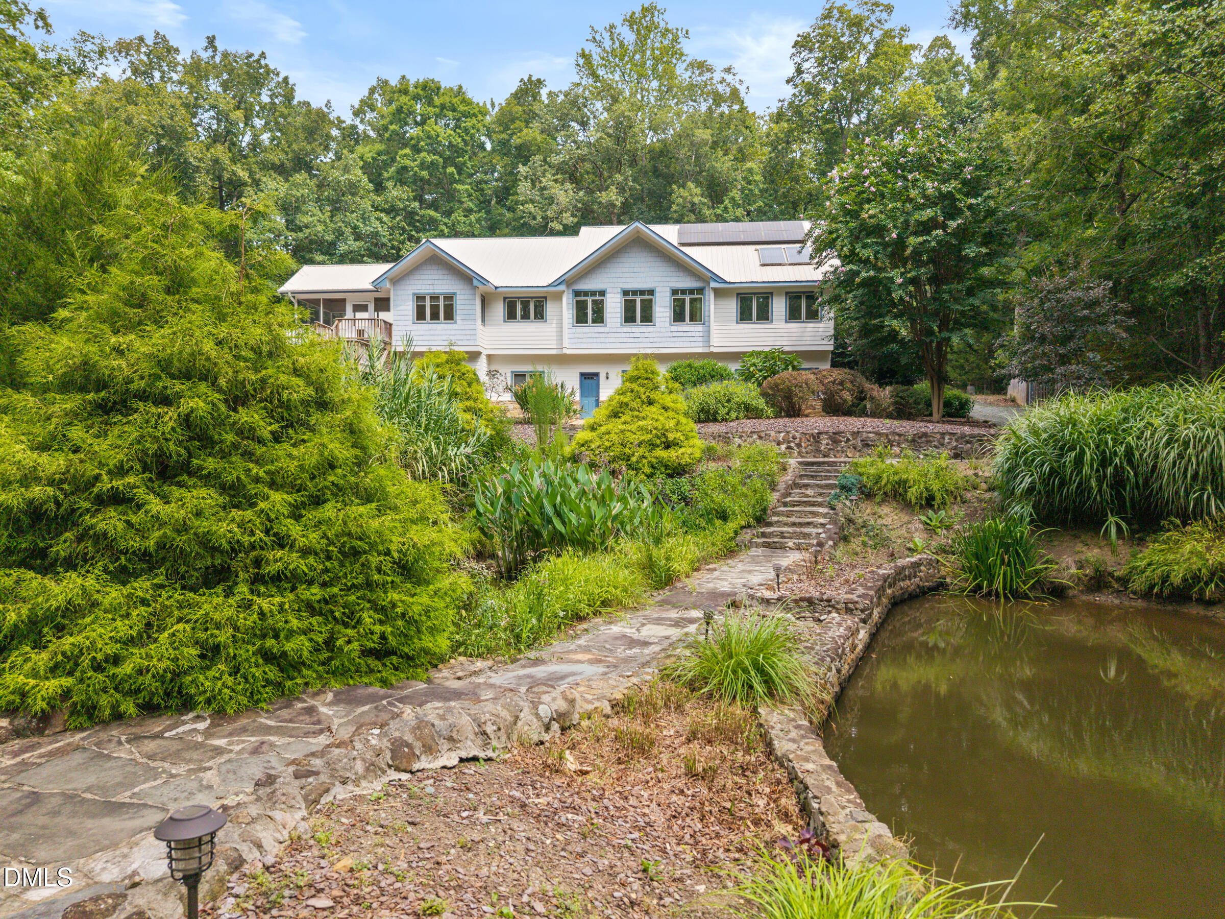 Light-colored home beyond a stone walkway and pond, surrounded by lush landscaping and mature trees.