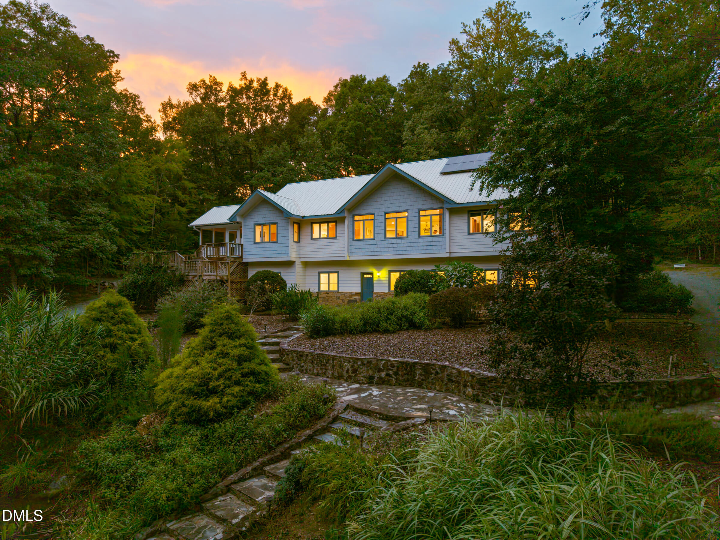 Wooded home with a metal roof, glowing windows, and stone garden steps at dusk.