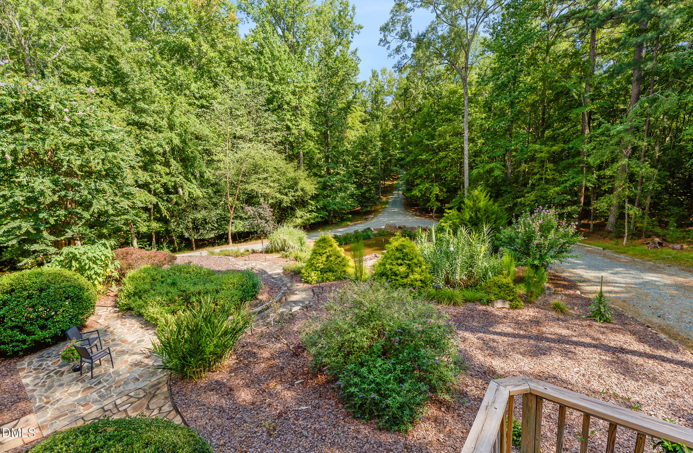 Wooded landscaped yard with a small pond, stone patio, and winding gravel drive.
