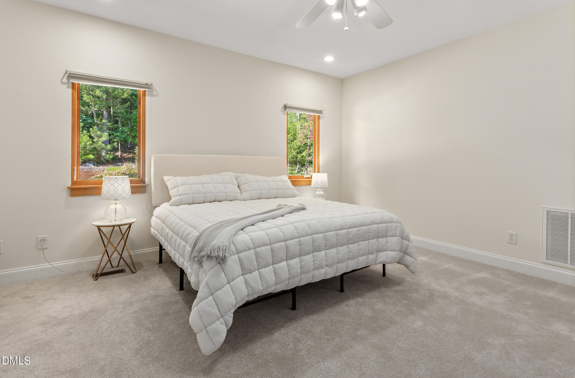 Staged bedroom with a white quilted bed between two wood-trimmed windows overlooking trees.