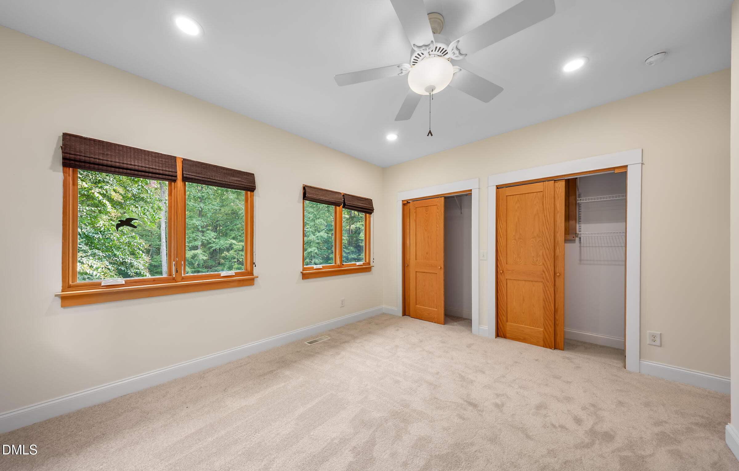 Empty carpeted bedroom with wood-trimmed windows overlooking trees and two closets.