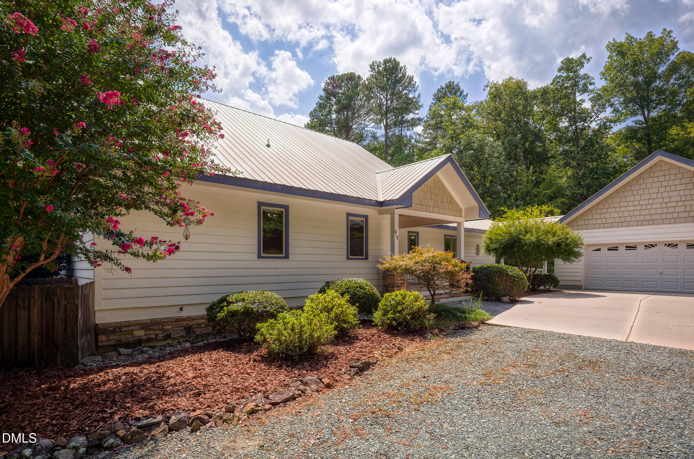 Light-colored home with a metal roof, covered porch, flowering crepe myrtle, and attached two-car garage.