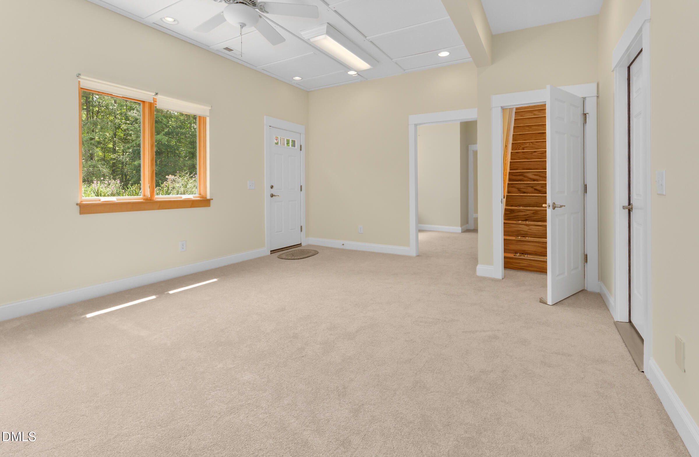 Empty carpeted room with a wood-trimmed window and an open doorway revealing a wooden staircase.