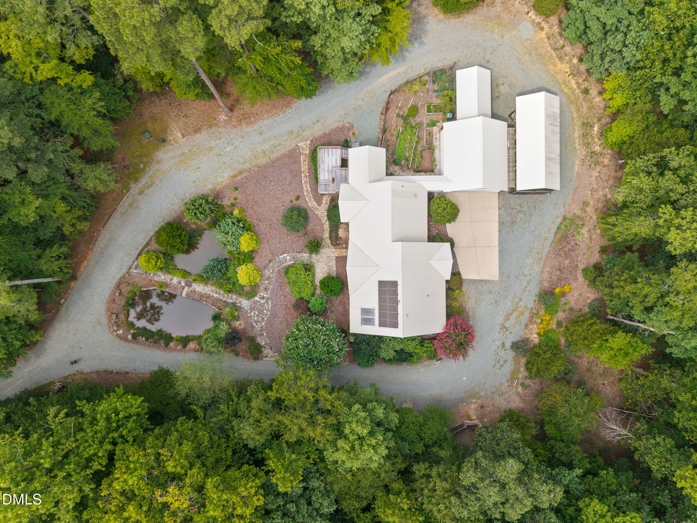 Aerial view of a wooded home with a looping gravel drive, landscaped gardens, ponds, and rooftop solar panels.