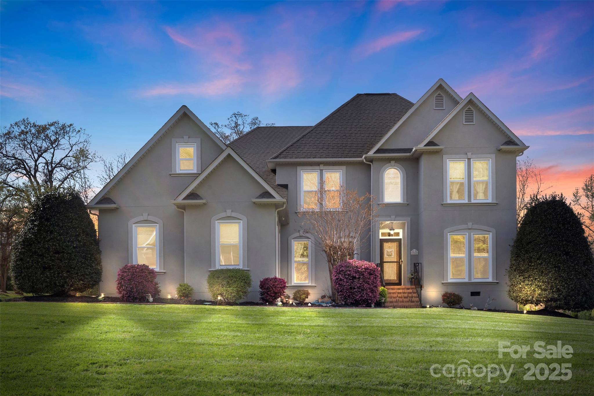 Gray two-story home with gabled roof and glowing windows at dusk, framed by a broad lawn and landscaped shrubs.