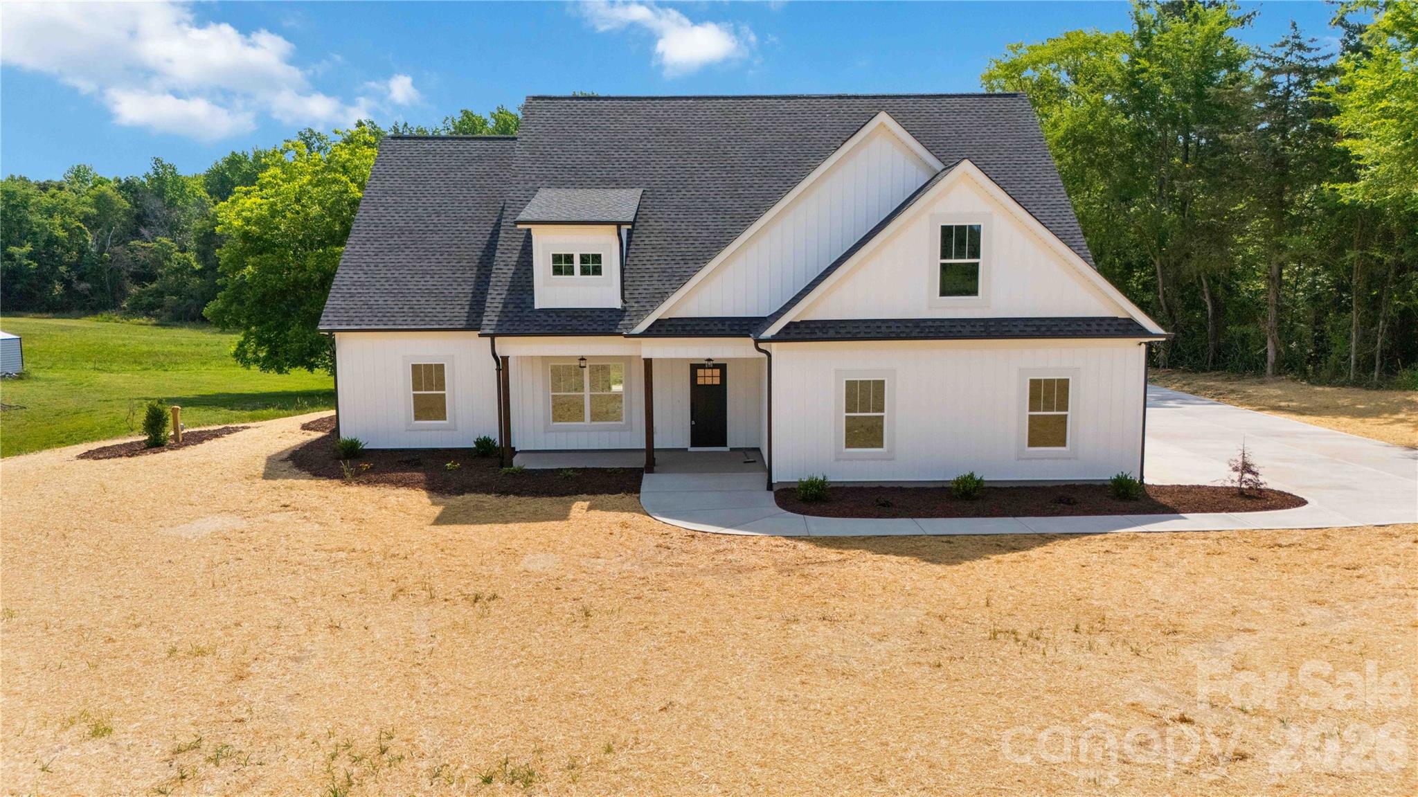 White farmhouse with a steep dark roof, front gables, and a covered entry on a wooded lot.