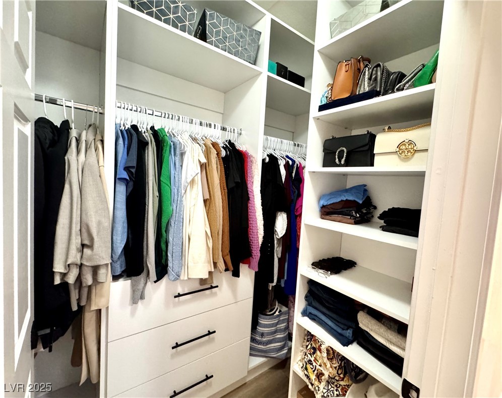 Walk-in closet with built-in white shelves, drawers, hanging clothes, and neatly arranged handbags.