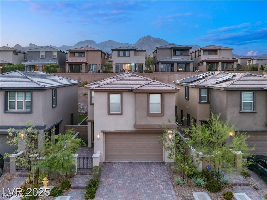 Two-story stucco home with an attached garage, paver driveway, and mountains visible behind neighboring houses.