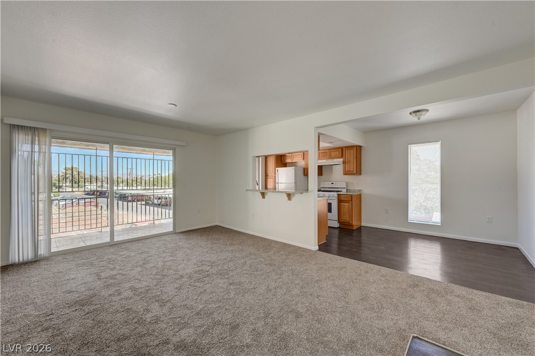 Empty living room with carpet, opening to a balcony and adjoining kitchen.