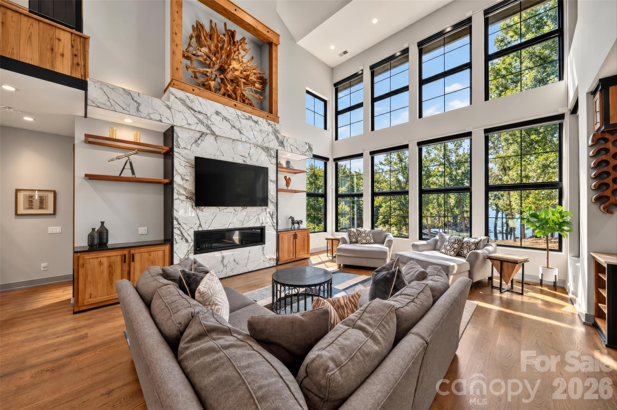 Double-height living room with a marble fireplace wall, wood cabinetry, and tall windows overlooking water.