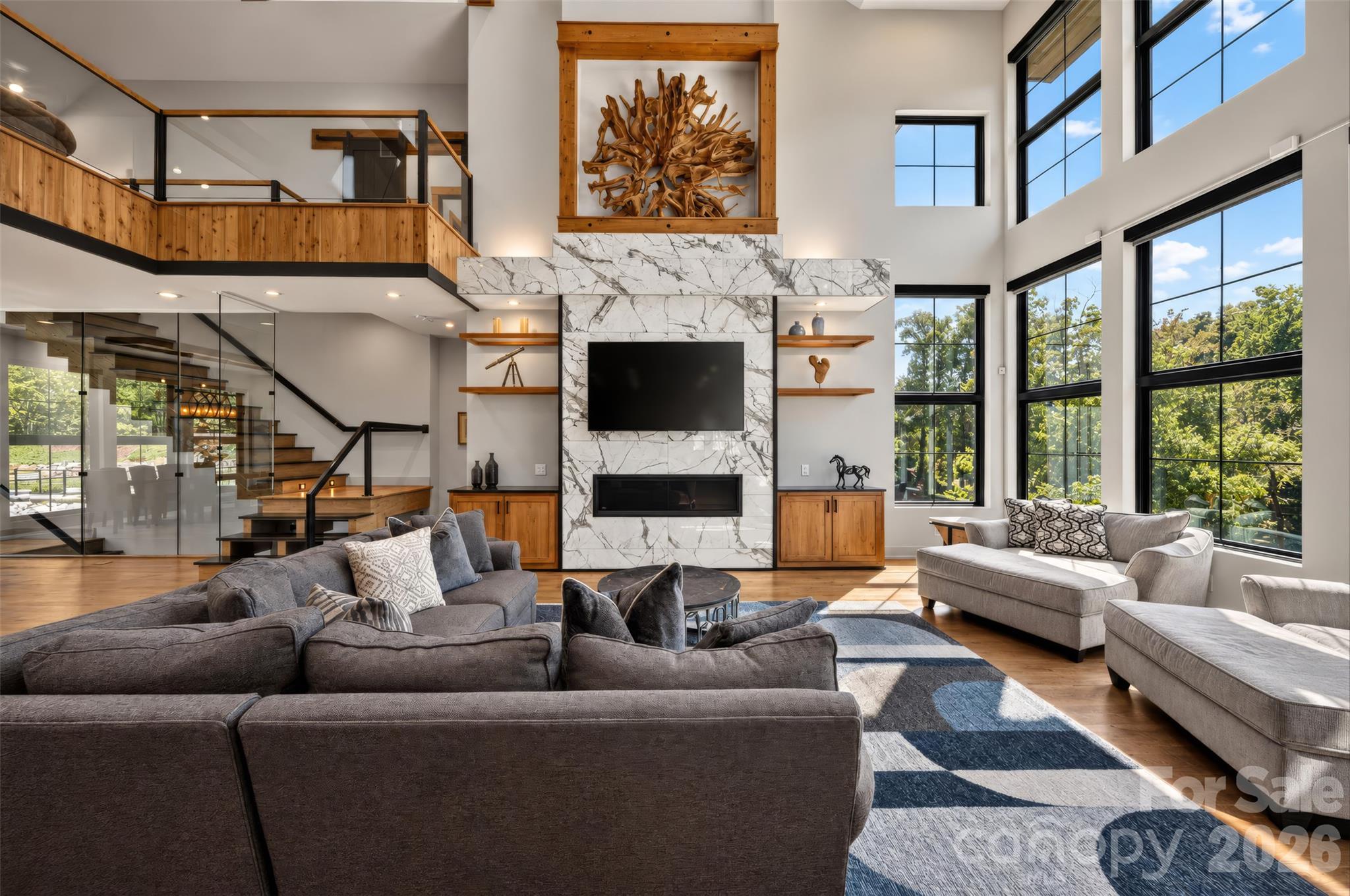 Double-height living room with a marble fireplace, glass-and-wood staircase, and tall windows overlooking trees.