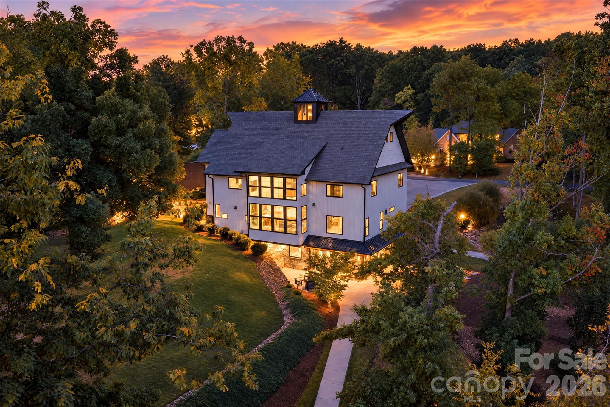 Modern white home with a dark gabled roof and warmly lit windows, surrounded by trees at dusk.